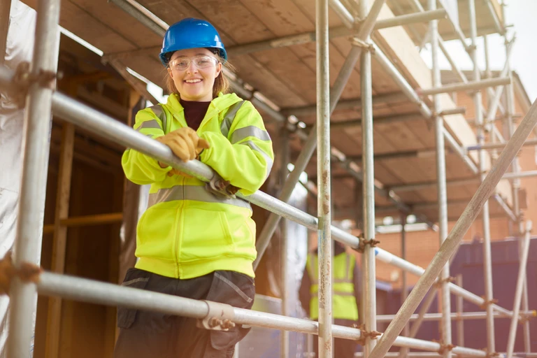 A female construction worker stands behind a scaffold and smiles to camera on a building site