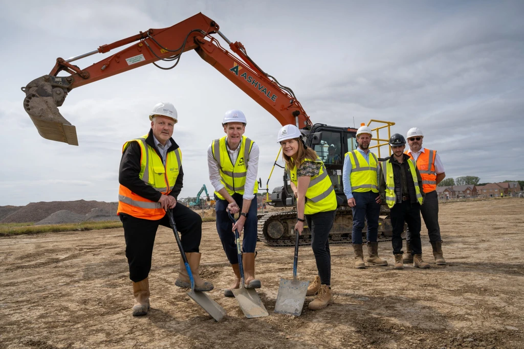Representatives from Amplius and Vistry in hard hats and hi vis vests breaking the ground at Wixams with a mechanical digger in the background