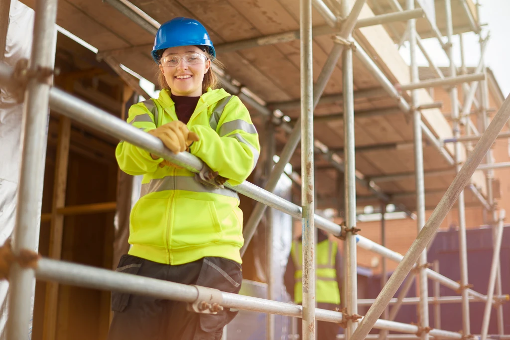 A female construction worker stands behind a scaffold and smiles to camera on a building site