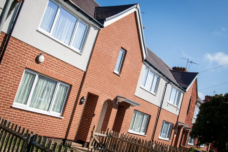 A row of modern two-storey terraced houses with red brick and light grey rendered exteriors. The homes feature white-framed windows, pitched dark tiled roofs, and small front gardens enclosed by low wooden picket fences. Front doors are set beneath sheltered porches, and the properties sit along a residential street under a clear blue sky on a sunny day.